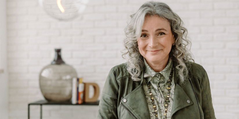Eine lächelnde Frau mit grauen lockigen Haaren trägt eine grüne Lederjacke und eine gemusterte Bluse. Im Hintergrund sieht man einen hellen Raum mit einem Regal und einer Vase.